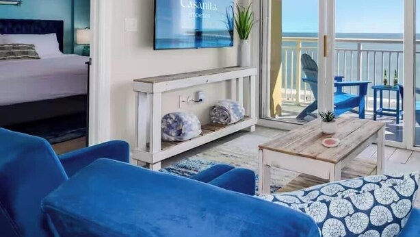 Ein Zimmer mit Bett, einem an der Wand befestigten Fernseher, einem Balkon mit Meerblick und blauen Möbeln.