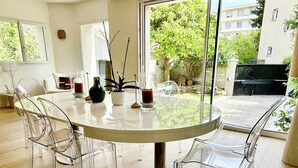 Un espace repas moderne avec une table ronde en verre, des chaises transparentes et une vue sur l’extérieur grâce à de grandes fenêtres.