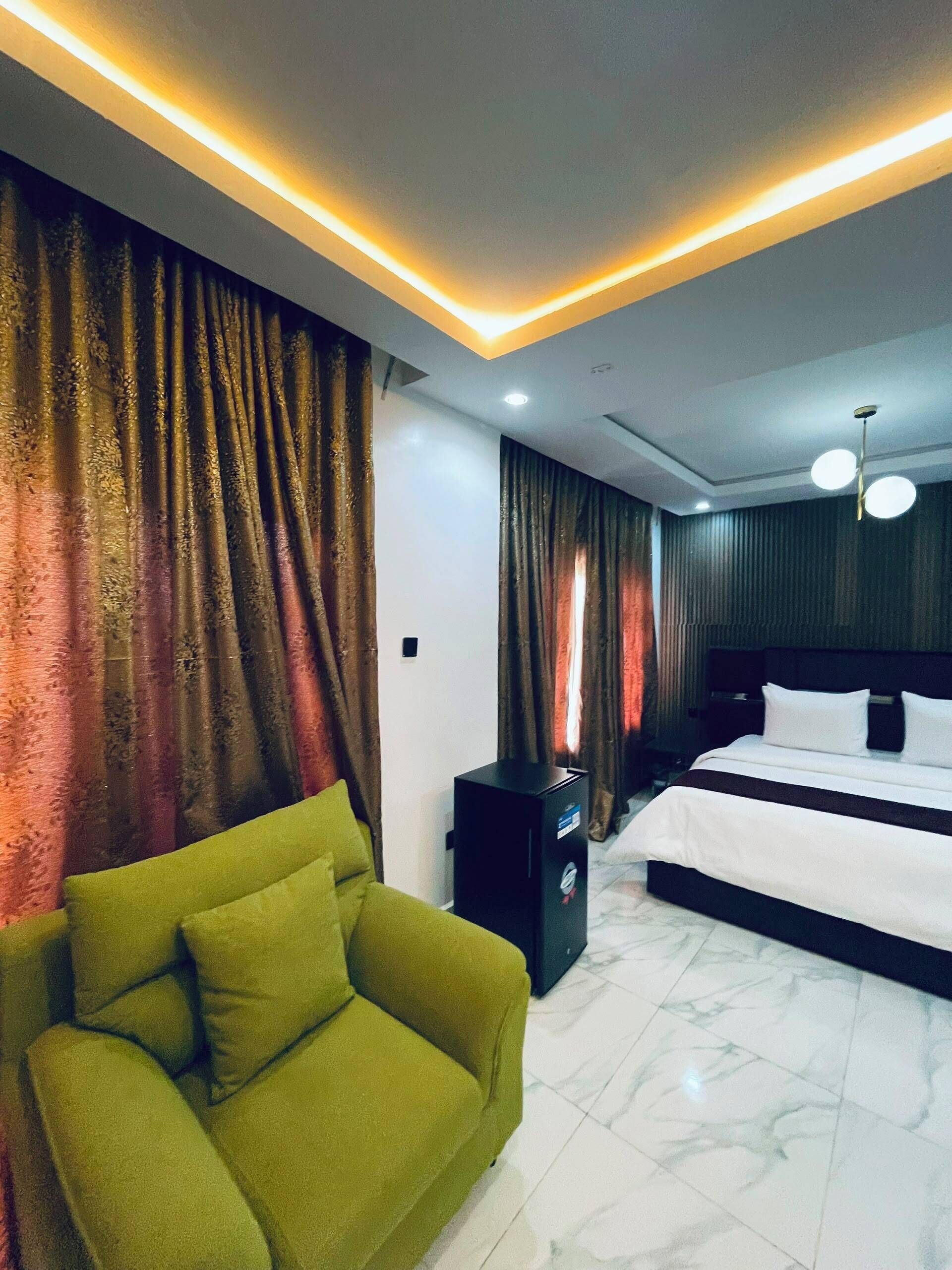 A modern hotel room with a bed, a green sofa, a TV stand, and a ceiling light fixture.