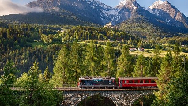 Ein Zug auf einer Brücke vor einer gebirgigen Landschaft.