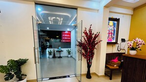 Área de recepção de hotel com entrada por porta de vidro, plantas em vasos, balcão de recepção e um quadro religioso emoldurado.