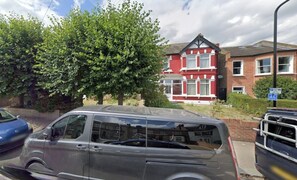 A residential street with parked cars, a red house with white trim, and a tree in the foreground. - Cosy 2BDR Flat in Leytonstone near Central Line (Greater London)