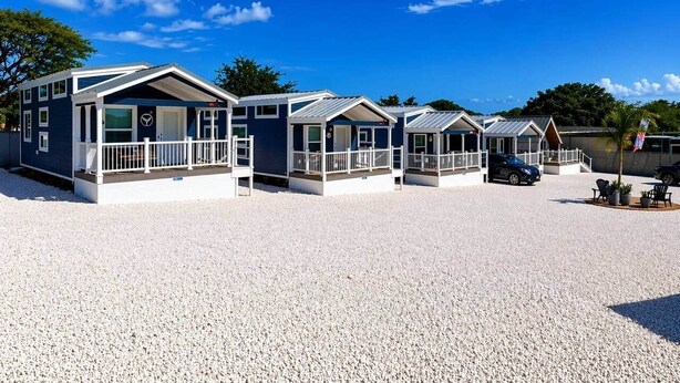 Une rangée de cabanes au bord de la plage, avec un sol de gravier, un ciel bleu dégagé et un drapeau.