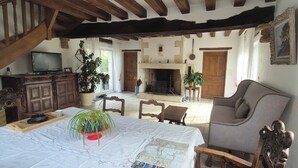 Un interior rústico con vigas de madera a la vista, una chimenea, una zona de comedor con mesa y sillas, y un televisor sobre un mueble.