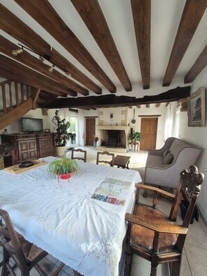 Ein rustikales Interieur mit freiliegenden Holzbalken, einem Kamin, einer Essecke mit Tisch und Stühlen sowie einem Fernseher auf einer Kommode.