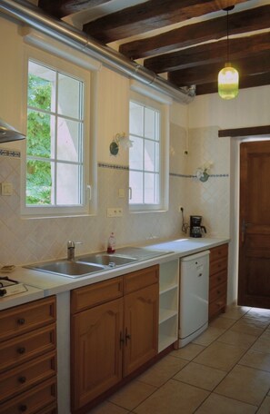 Eine Küche mit Holzschränken, einer weißen Arbeitsplatte, einem Spülbecken und einem Geschirrspüler. Zwei Fenster sorgen für natürliches Licht und eine Hängelampe ist angebracht.