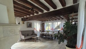 Una habitación amplia con techo alto, vigas de madera, piso de baldosas, un sofá, una mesa de comedor y una planta grande en maceta.