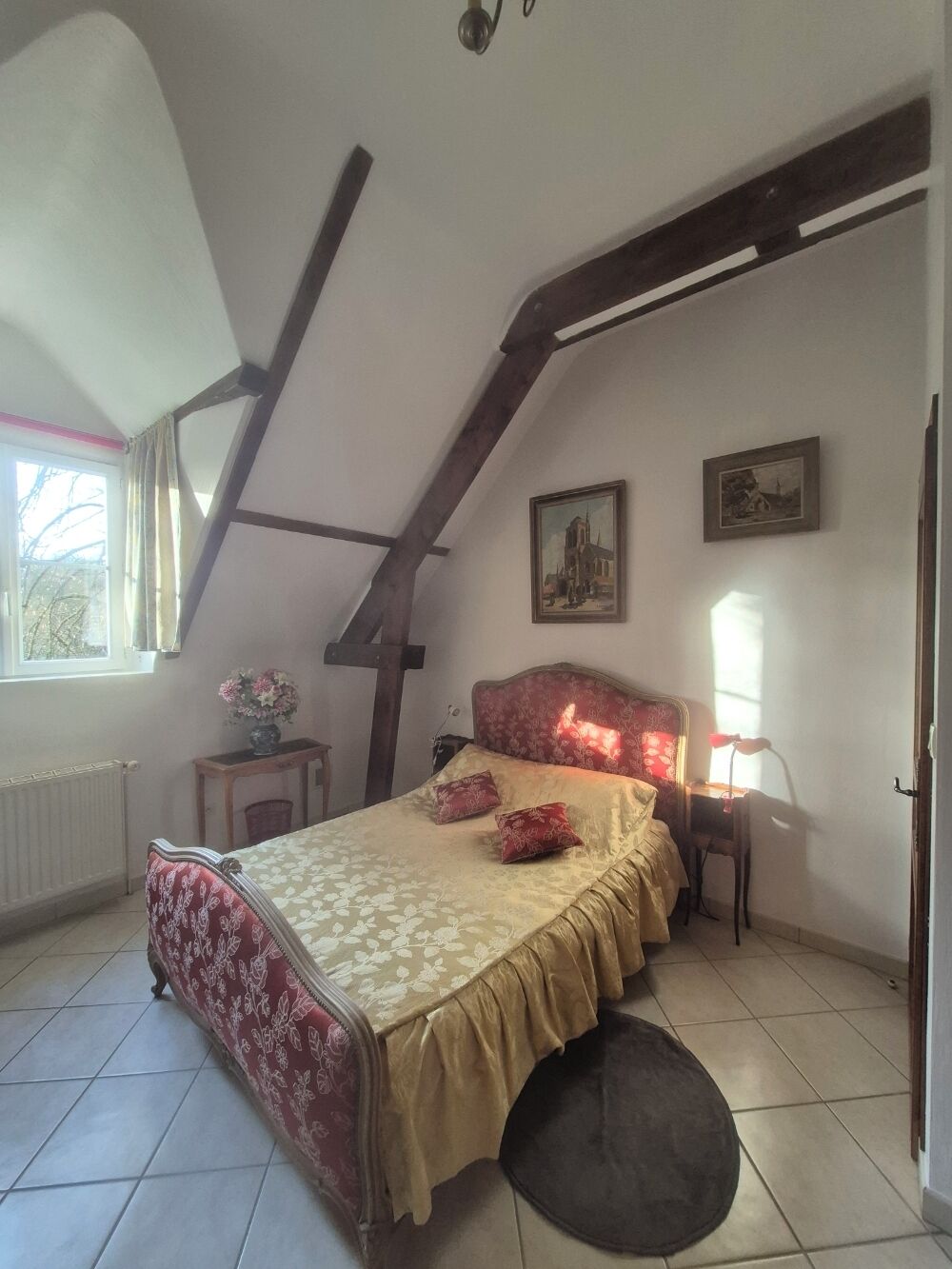 A bedroom with a sloped ceiling, a bed with a floral bedspread, a small table with a vase, and a window with curtains.