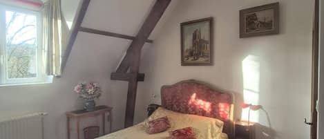 A bedroom with a sloped ceiling, a bed with a floral bedspread, a small table with a vase, and a window with curtains.