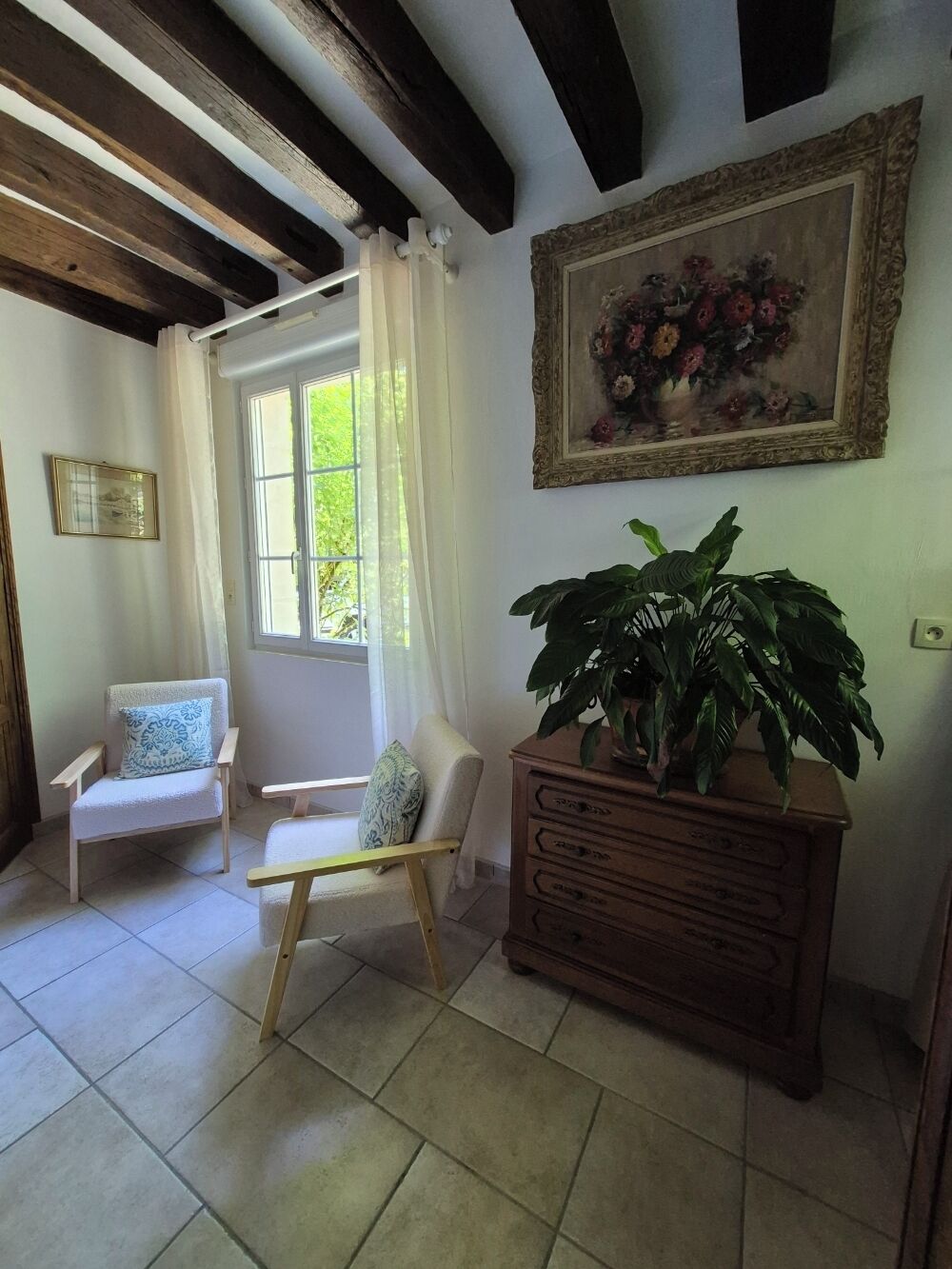 A room with a wooden ceiling, a window with curtains, a chair, a wooden dresser, and a painting on the wall.