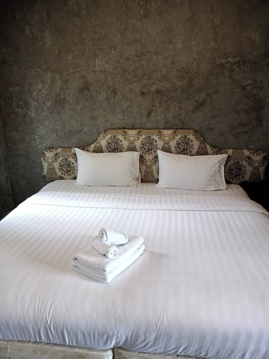 Una cama bien hecha con sábanas y almohadas blancas, un cabecero con estampado floral y una pila de toallas dobladas sobre la cama.