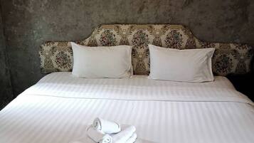 Una cama bien hecha con sábanas y almohadas blancas, un cabecero con estampado floral y una pila de toallas dobladas sobre la cama.