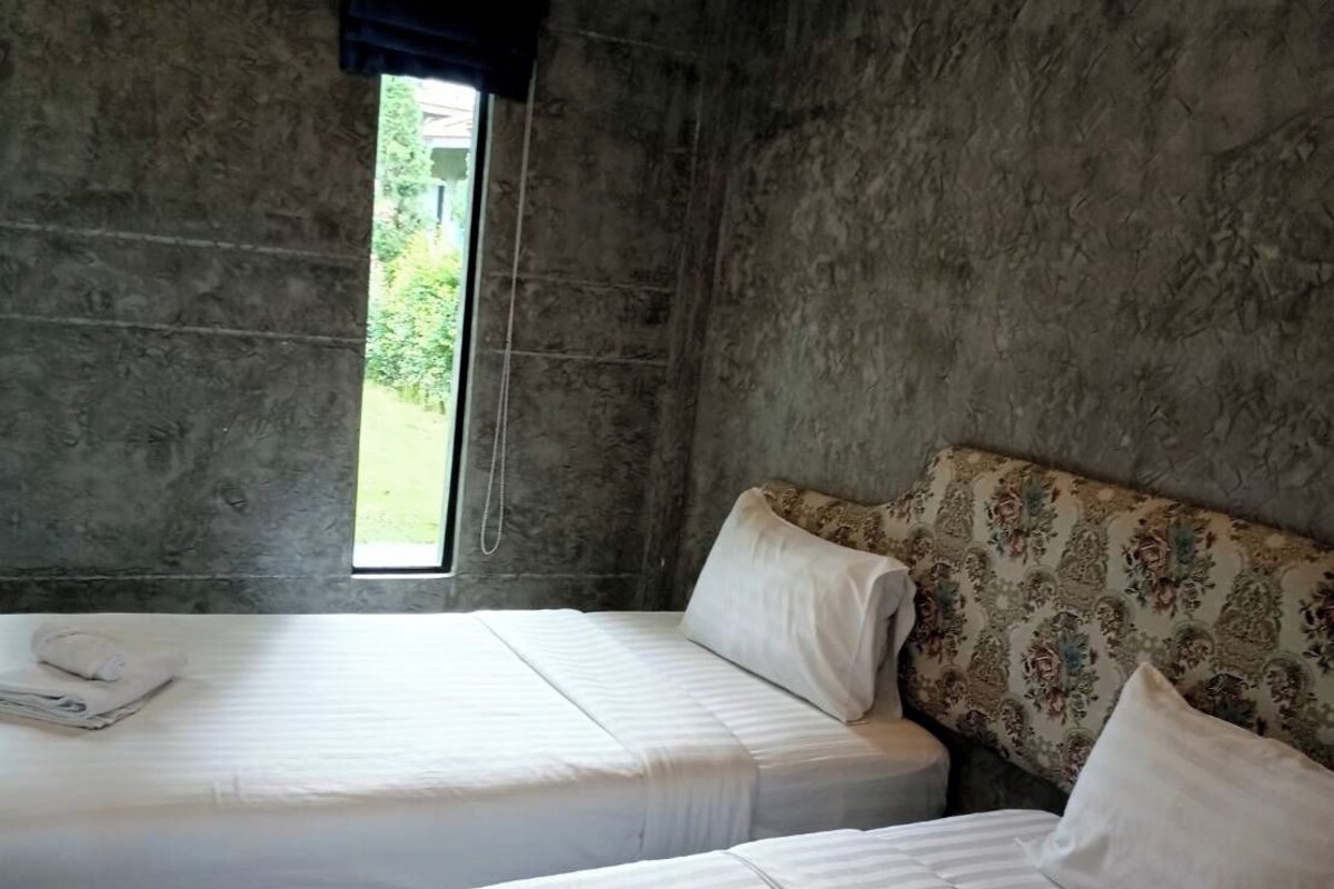 A room with two beds, each with white linens and pillows, a window with a view of greenery, and a textured wall.