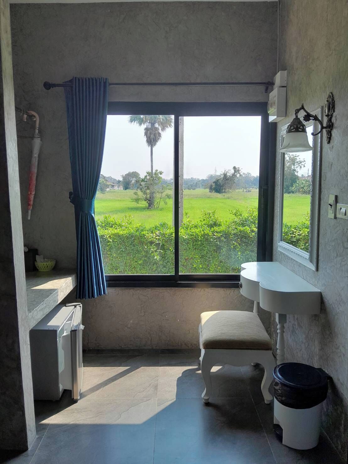 A room with a large window, a white sink, a bench, and a trash bin.