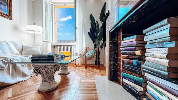 Quarto com sofá, mesa de vidro, estante, planta e vista do céu pela janela.