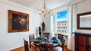 Uma sala de jantar com uma mesa redonda posta para seis pessoas, rodeada por cadeiras de madeira, um candelabro e um quadro grande emoldurado na parede.