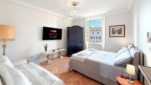 Quarto com cama, mesa de cabeceira com abajur, TV exibindo serviços de streaming, janela com vista e guarda-roupa escuro.