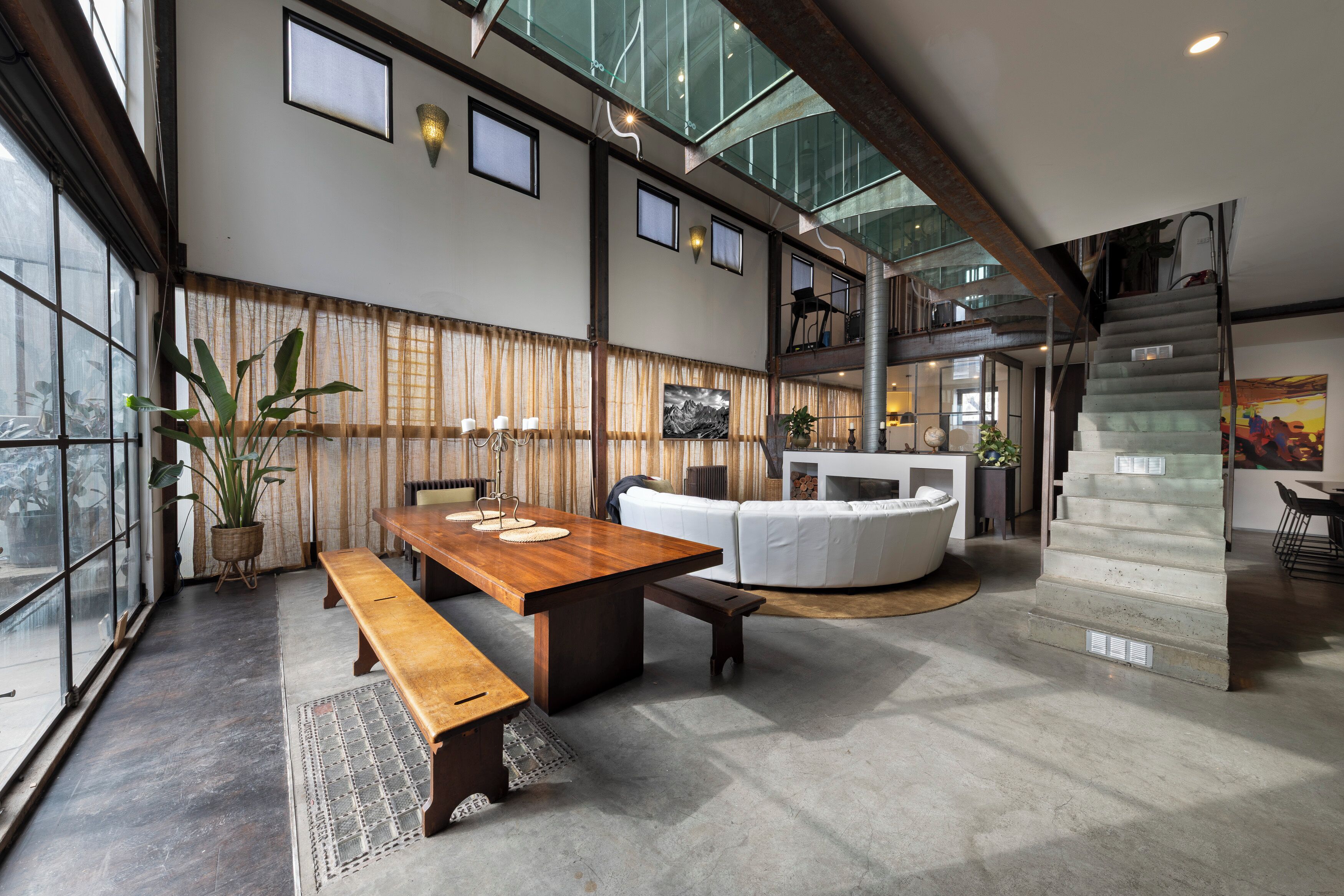 A spacious interior with a high ceiling, a wooden dining table, a white sofa, and a staircase leading to an upper level.