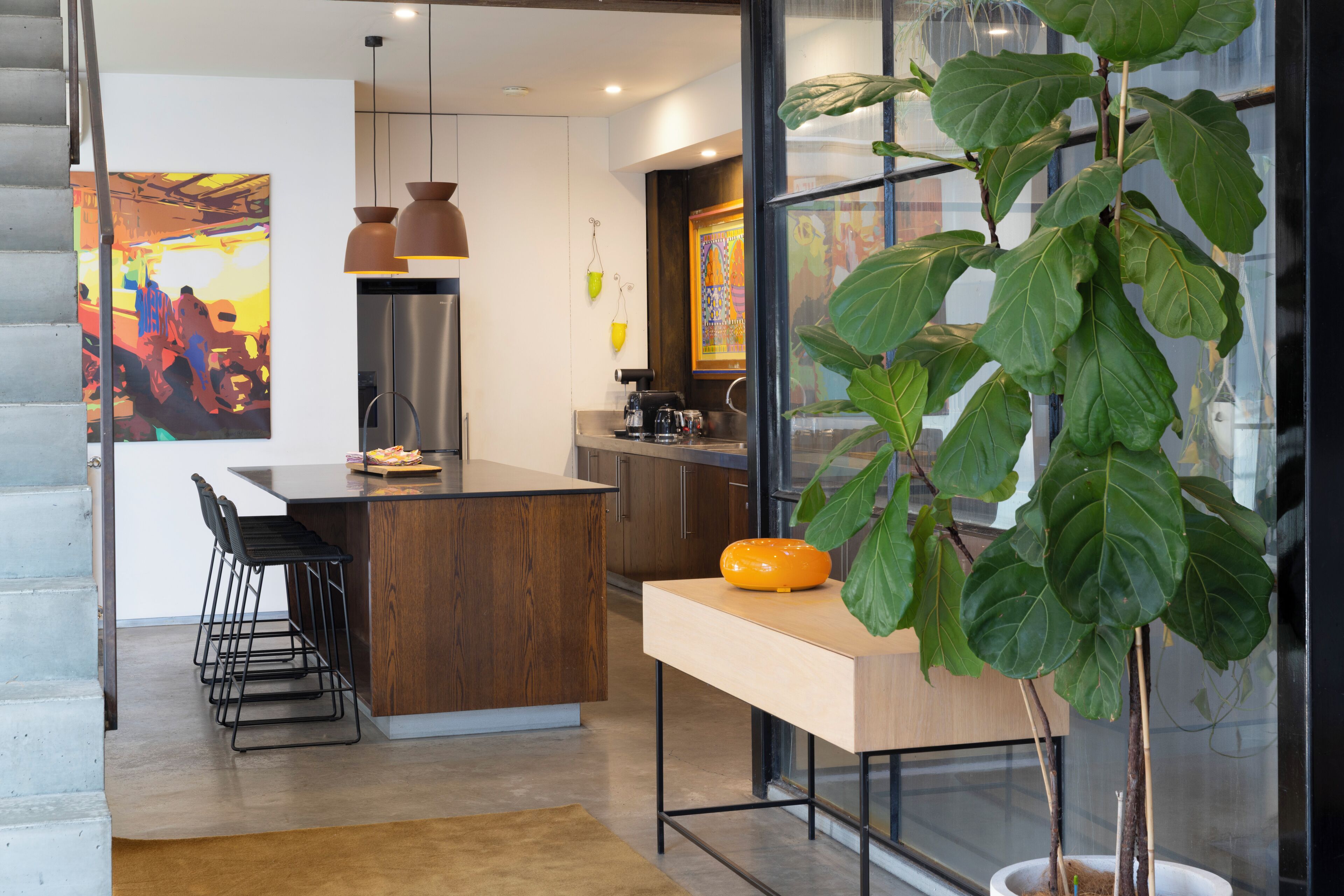 A modern kitchen with a central island, bar stools, pendant lighting, and a large green plant.