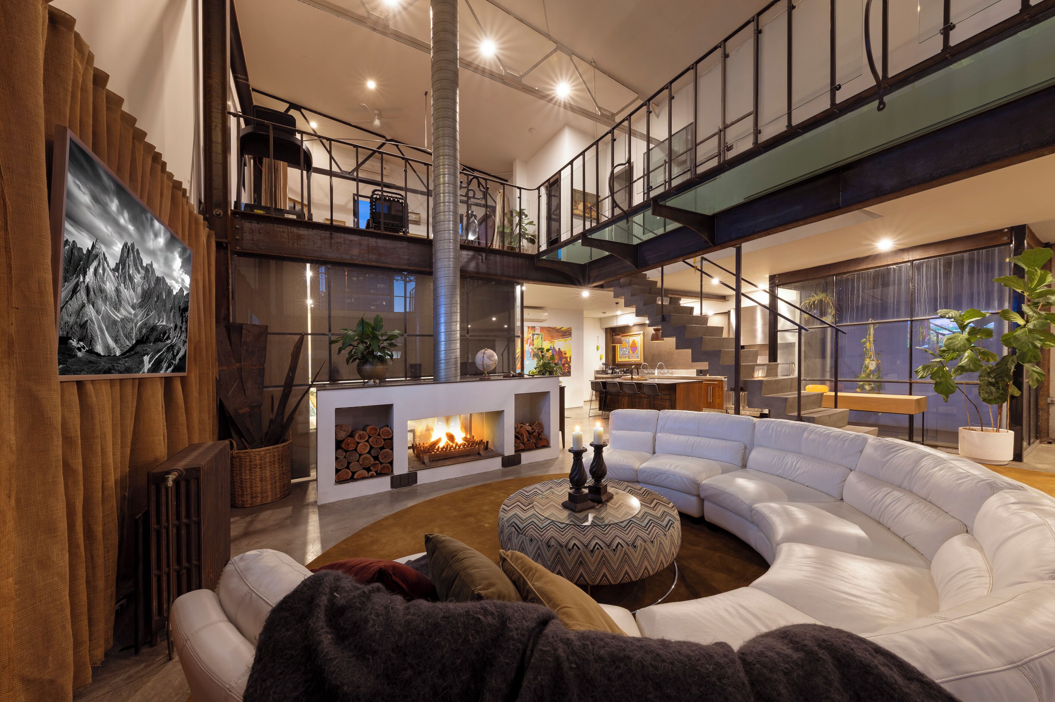 A modern living room with a curved white sofa, a fireplace, a staircase, and a dining area.