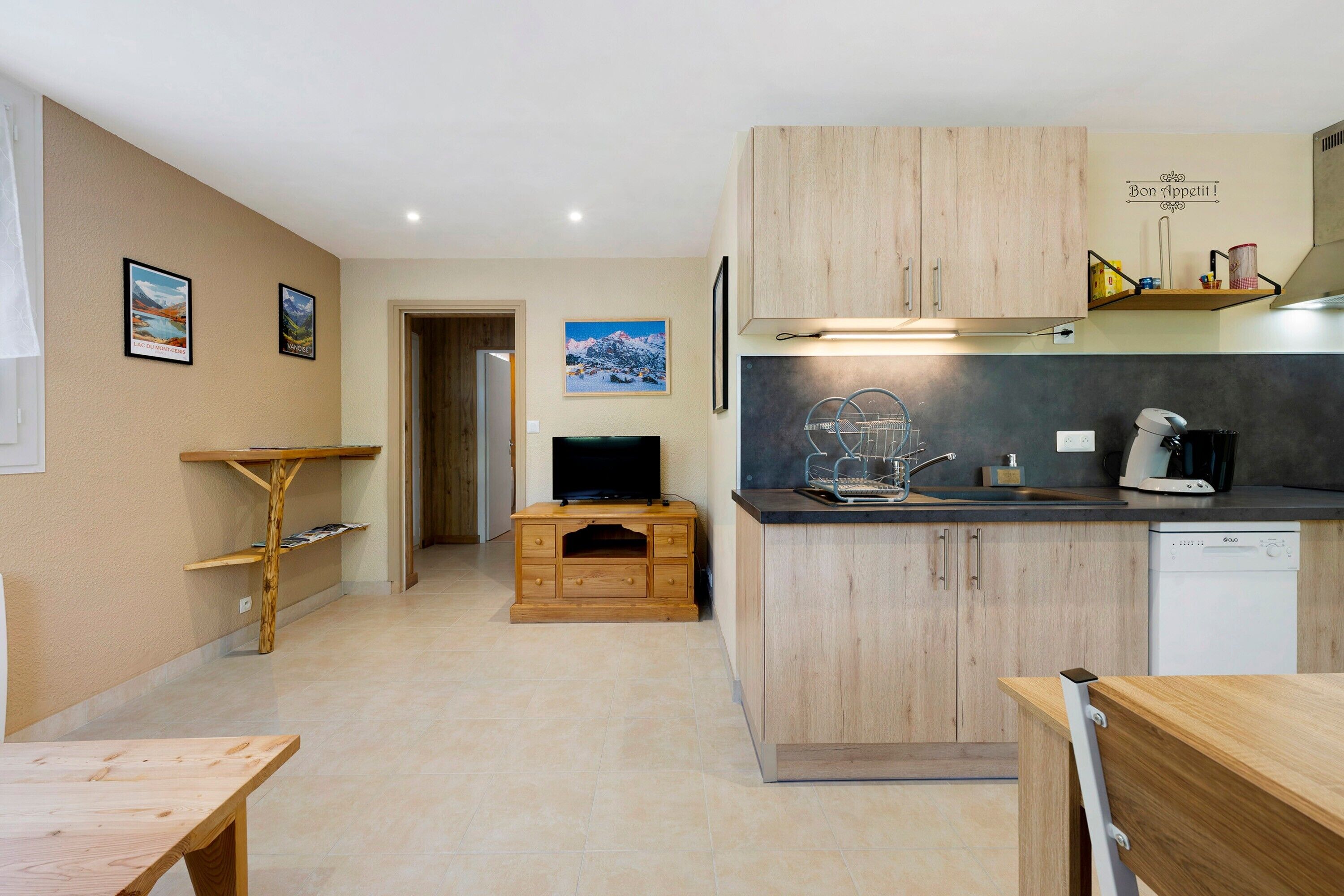 Een kleine keuken met houten keukenkastjes, een inbouw vaatwasser en een eethoek met een houten tafel.