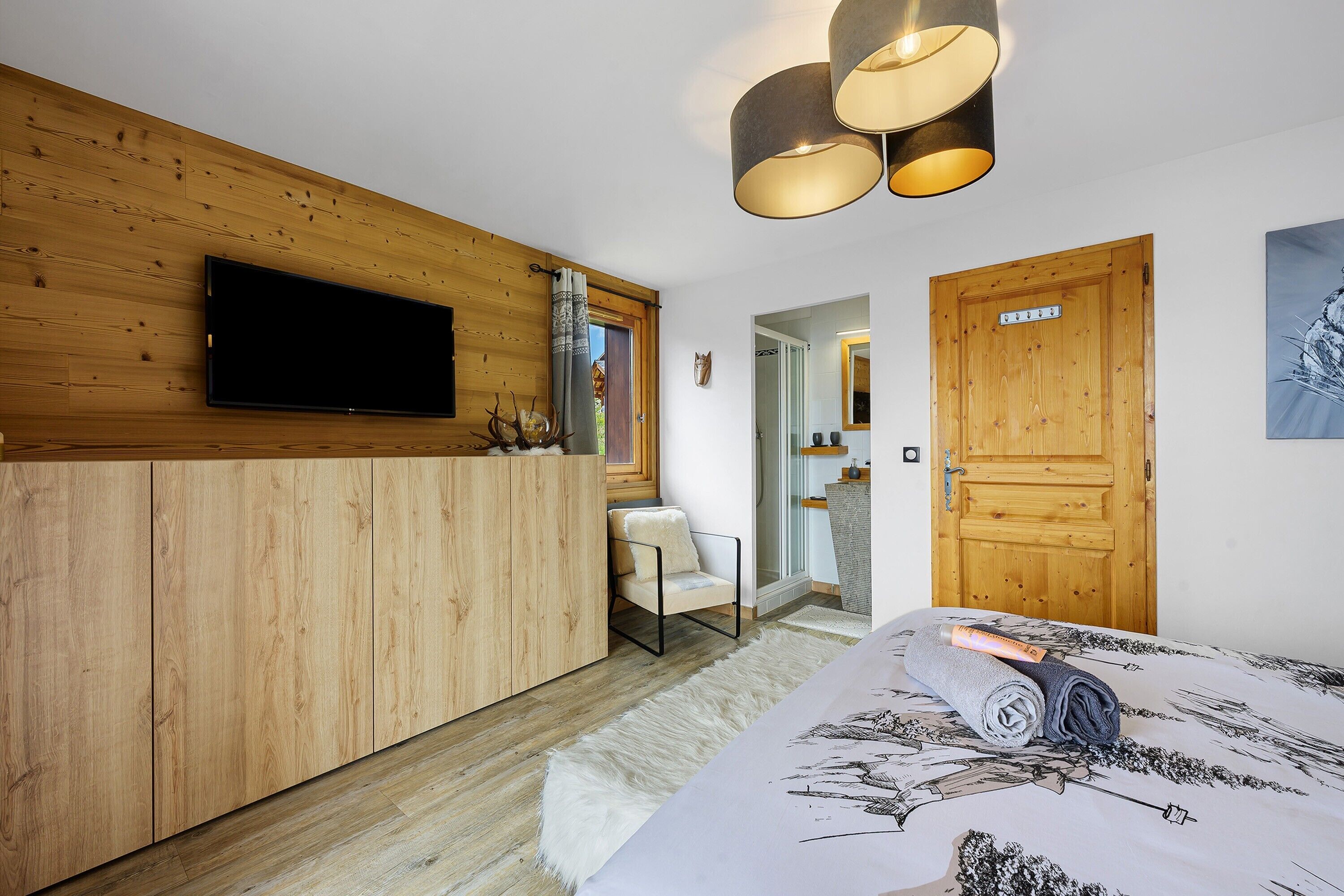 A bedroom with a wooden wardrobe, a bed with a patterned bedspread, a television mounted on the wall, and a door with a mirror.