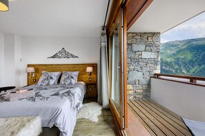 A bedroom with a wooden headboard, a bed with patterned bedding, a nightstand, and a view of a mountainous landscape through a balcony. - High-End Apartment in La Rosière 1850, 5 Rooms for 10 Guests, Ski-In/Ski-Out (Montvalezan)