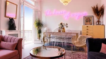 A modern living room with a glass coffee table, a pink sofa, a chandelier, and a neon sign reading 'The house of vogue'.