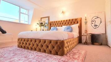 A bedroom with a tufted headboard, a pink patterned rug, a bedside table with a lamp, and a wall-mounted light fixture.