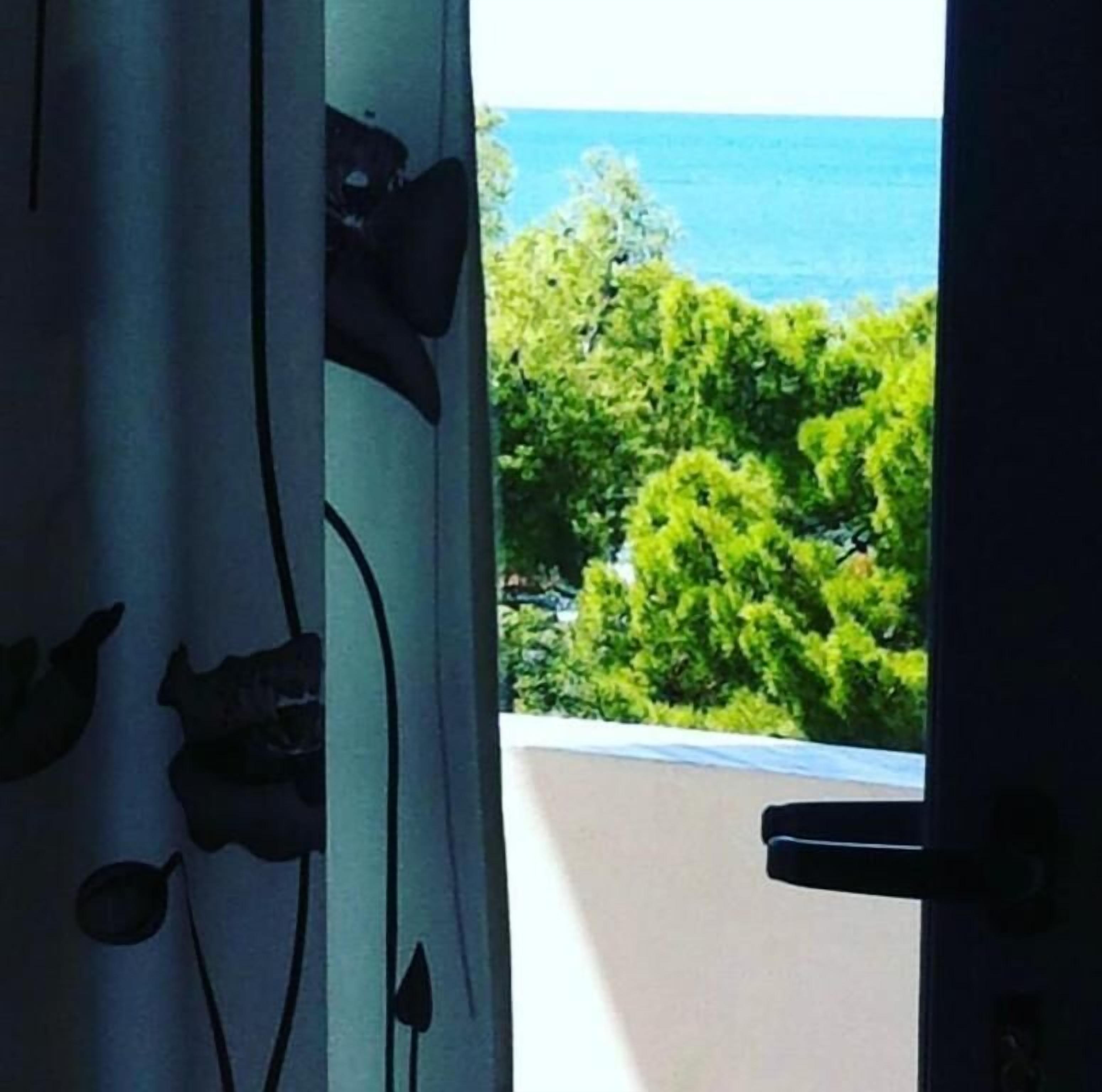 A view through a window with a curtain featuring a floral pattern, a balcony, and a body of water in the background.