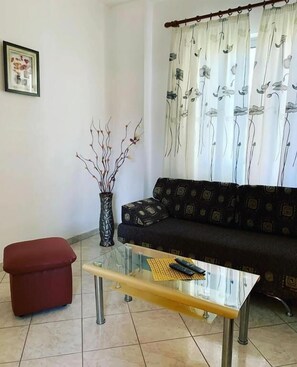Uma sala de estar com sofá, mesa de centro de vidro, um banquinho vermelho, um vaso com galhos e uma cortina estampada com flores.