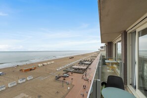 Vista a la playa desde un balcón con una mesa y sillas, una playa de arena y sombrillas.