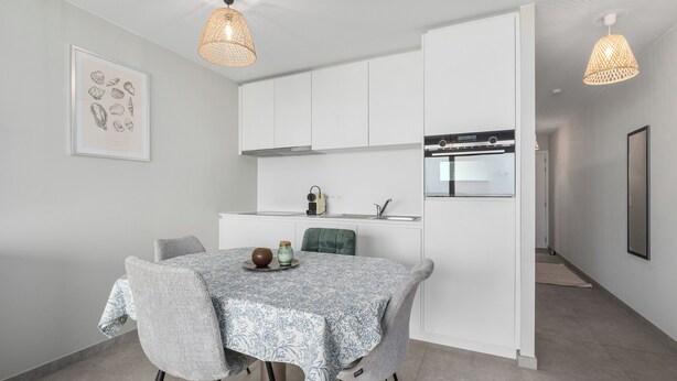 A modern kitchen with white cabinets, a dining table with a patterned tablecloth, and pendant lights. - 1 bedroom accommodation in De Panne (De Panne)