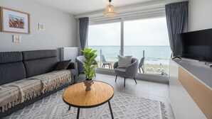 A modern living room with a sofa, armchair, TV, and a view of the beach. - 1 bedroom accommodation in De Panne (De Panne)