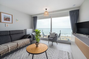 A modern living room with a sofa, armchair, TV, and a view of the beach. - 1 bedroom accommodation in De Panne (De Panne)