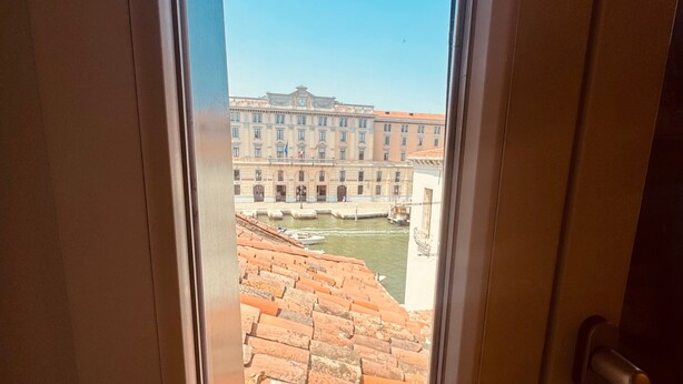 Vista a través de una ventana que muestra un edificio con tejado de tejas y una construcción grande al fondo.