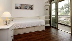 Une chambre à coucher comprenant un lit, une table de chevet avec une lampe, une étagère fixée au mur et une porte-fenêtre coulissante donnant sur un balcon.