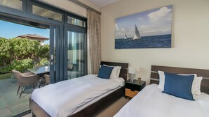 Ein Hotelzimmer mit zwei Betten, einer Glastür, die auf eine Terrasse führt, einem Segelboot-Gemälde und einem Nachttisch mit einer Lampe.