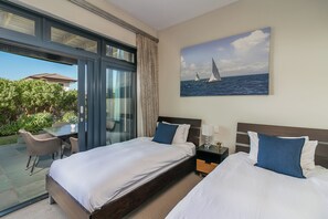 Ein Hotelzimmer mit zwei Betten, einer Glastür, die auf eine Terrasse führt, einem Segelboot-Gemälde und einem Nachttisch mit einer Lampe.