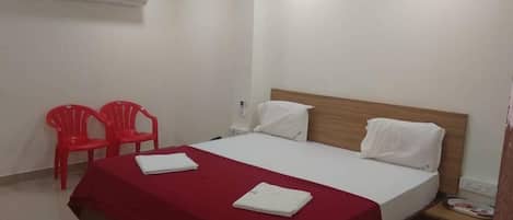 A hotel room with a bed, two red chairs, a nightstand, and an air conditioning unit.