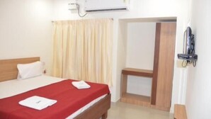 Habitación de hotel con cama, cabecera de madera, colcha roja, almohada blanca, aire acondicionado de pared, cortina y puerta que da a otra habitación.
