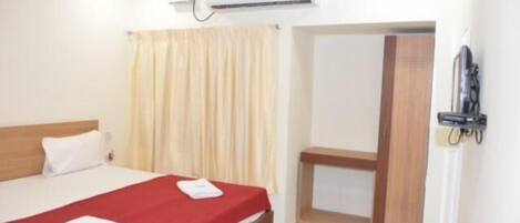 Habitación de hotel con cama, cabecera de madera, colcha roja, almohada blanca, aire acondicionado de pared, cortina y puerta que da a otra habitación.