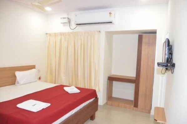 A hotel room with a bed, a wooden headboard, a red bedspread, a white pillow, a wall-mounted air conditioner, a curtain, and a door leading to another room.