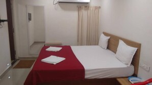 Una habitación de hotel con cama, colcha roja, almohada blanca, cabecera de madera, mesita de noche y un equipo de aire acondicionado.
