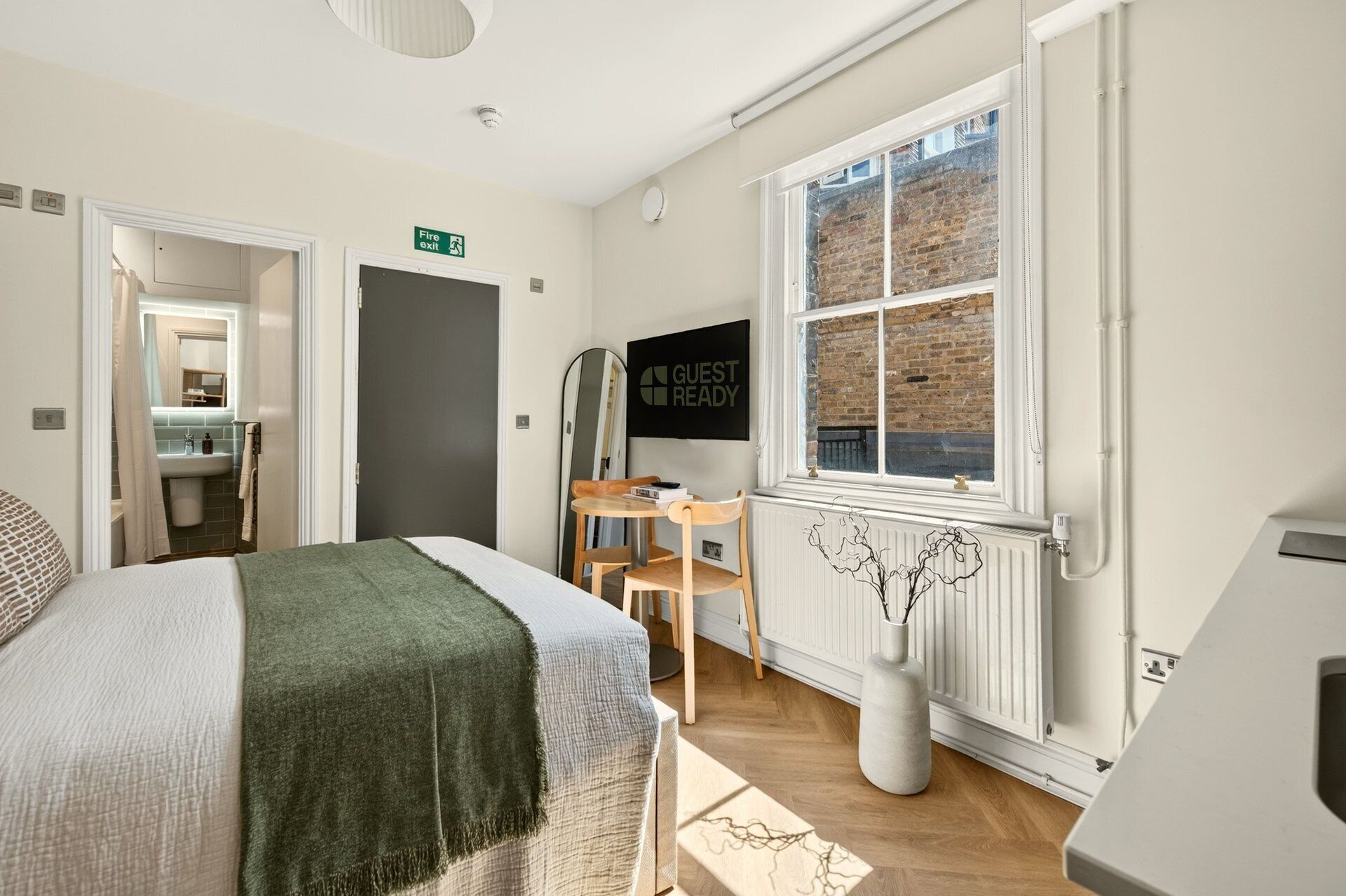 A modern hotel room with a bed, a desk, a chair, a TV, a window with a view of a brick building, and a fire exit sign.