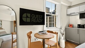 Een moderne kamer met een bed, een klein rond tafeltje met stoelen, een bordje 'GUEST READY', een raam en een kitchenette.