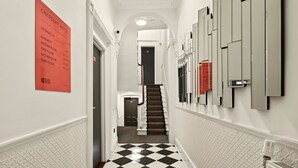 Een hotelgang met een geruit vloertapijt, een trap en een bord aan de muur met kamer- en verdiepingnummers.