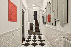 A hotel hallway with a checkered floor, a staircase, and a wall-mounted sign indicating room numbers and floors. - GuestReady - Stylish Double En-Suite in London (London)