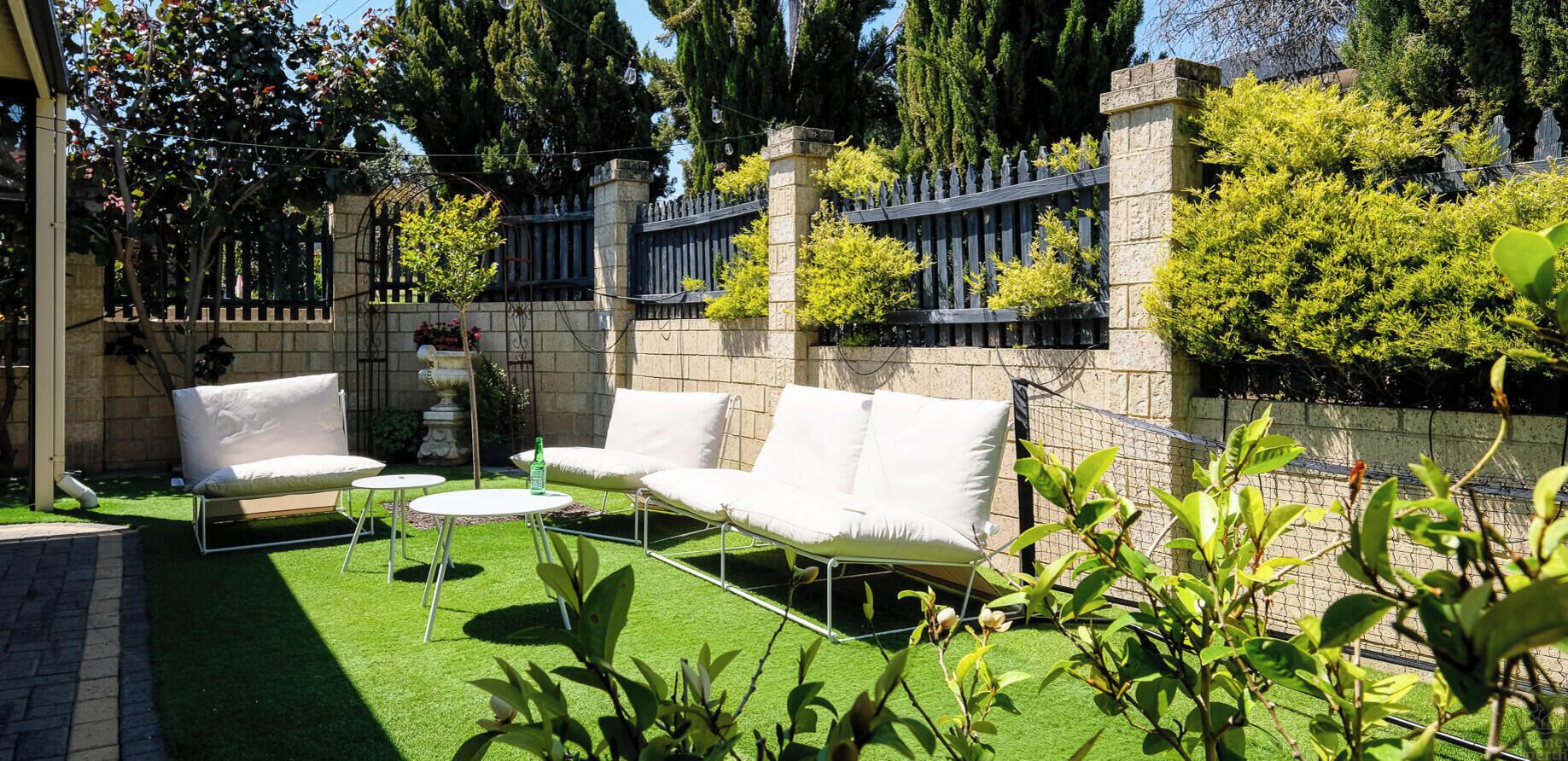 An outdoor seating area with a white sofa and two chairs, surrounded by a stone wall and greenery.