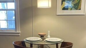 A dining area with a round glass table, two chairs, a hanging light fixture, a framed artwork, and a window with blinds. - Cozy Coach House: just minutes to CMU and Soaring Eagle Casino (Mount Pleasant)